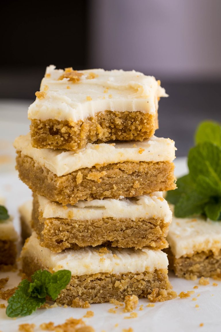 A stack of four frosted cookie bars with crumbs scattered around, garnished with a fresh mint sprig.