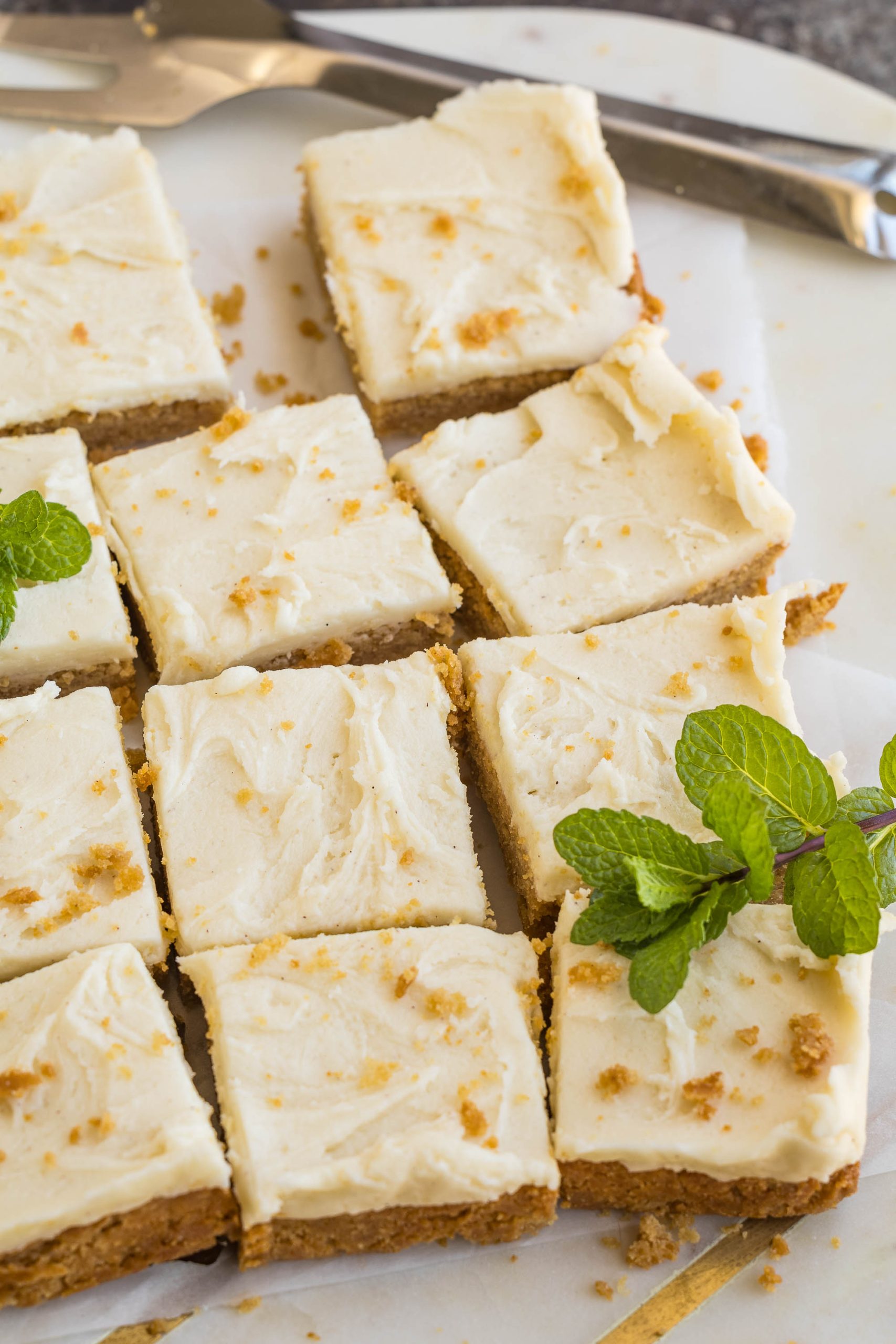 Sliced cheesecake squares with a crumbly base, topped with creamy frosting, garnished with fresh mint leaves on parchment paper.