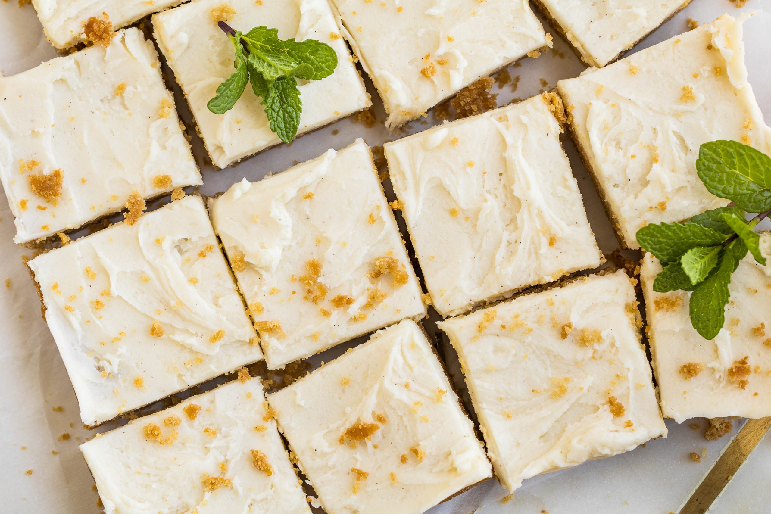 Squares of peanut butter graham cracker blondies topped with crumbled pieces and garnished with fresh mint leaves are arranged in a grid on a white surface.