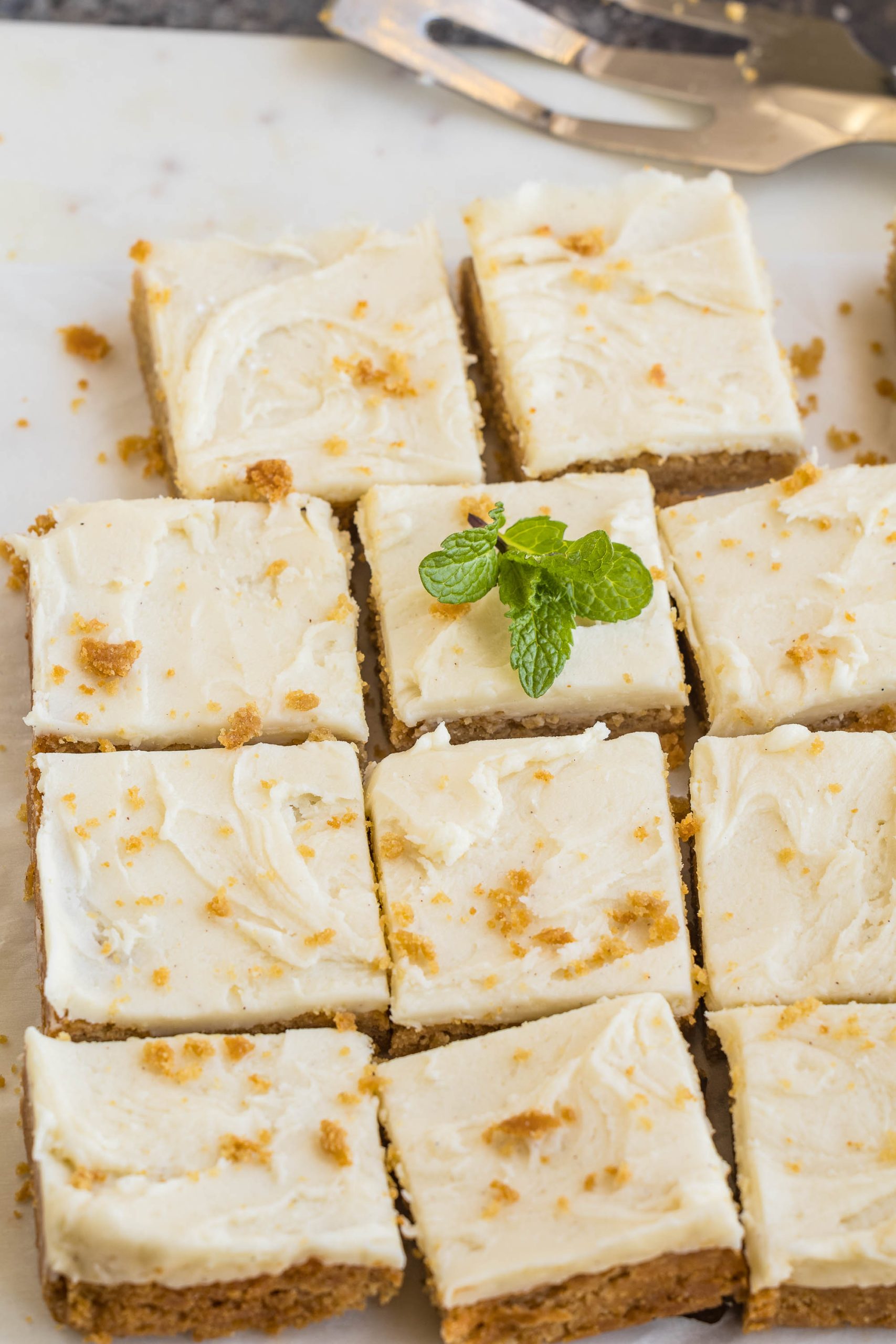 Nine square cheesecake bars with creamy frosting and crumb topping, arranged on a surface. One bar is garnished with a mint sprig.