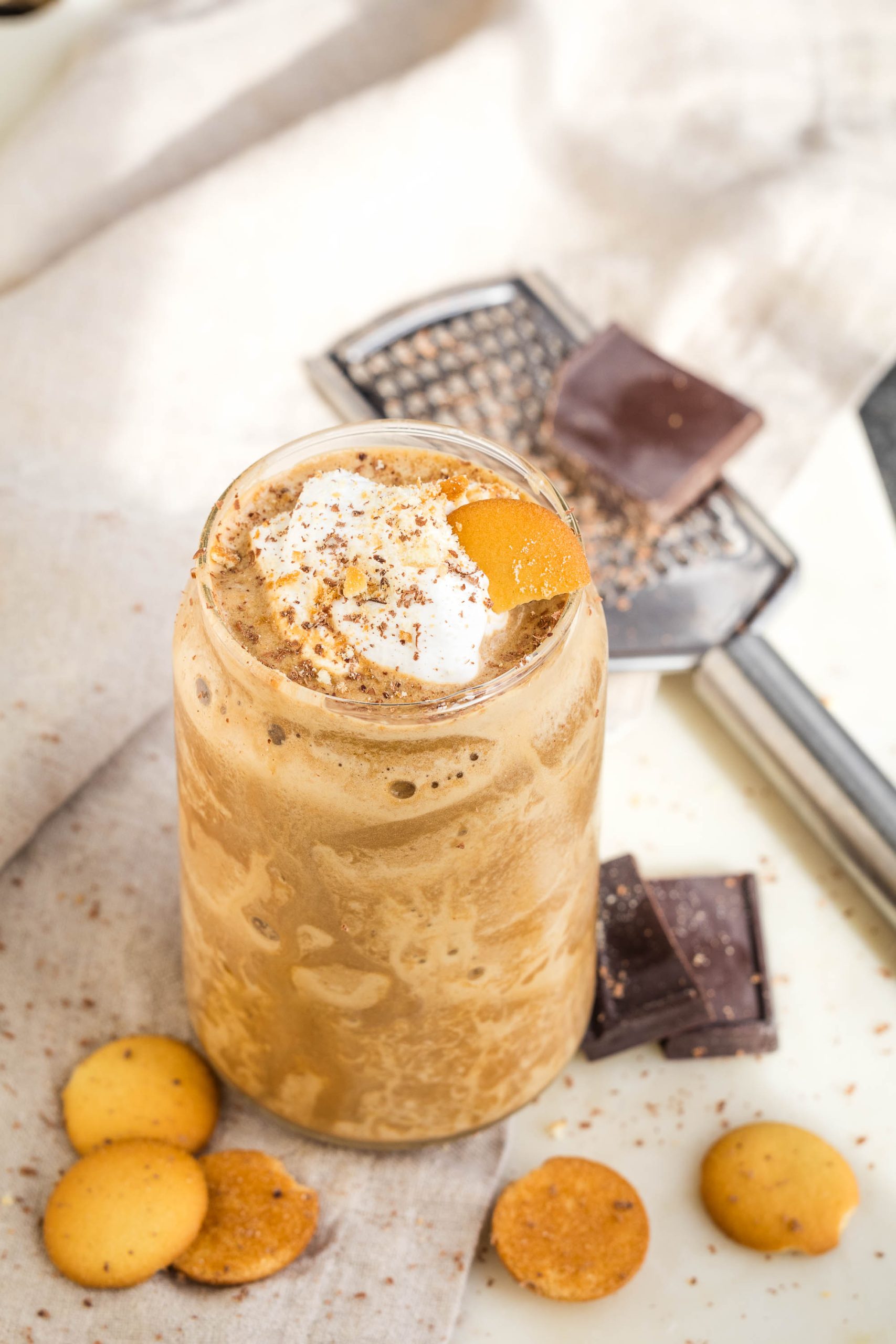 A tall glass of Protein Tiramisu Smoothie topped with whipped cream and a citrus peel, surrounded by chocolate pieces and round cookies on a light cloth surface.