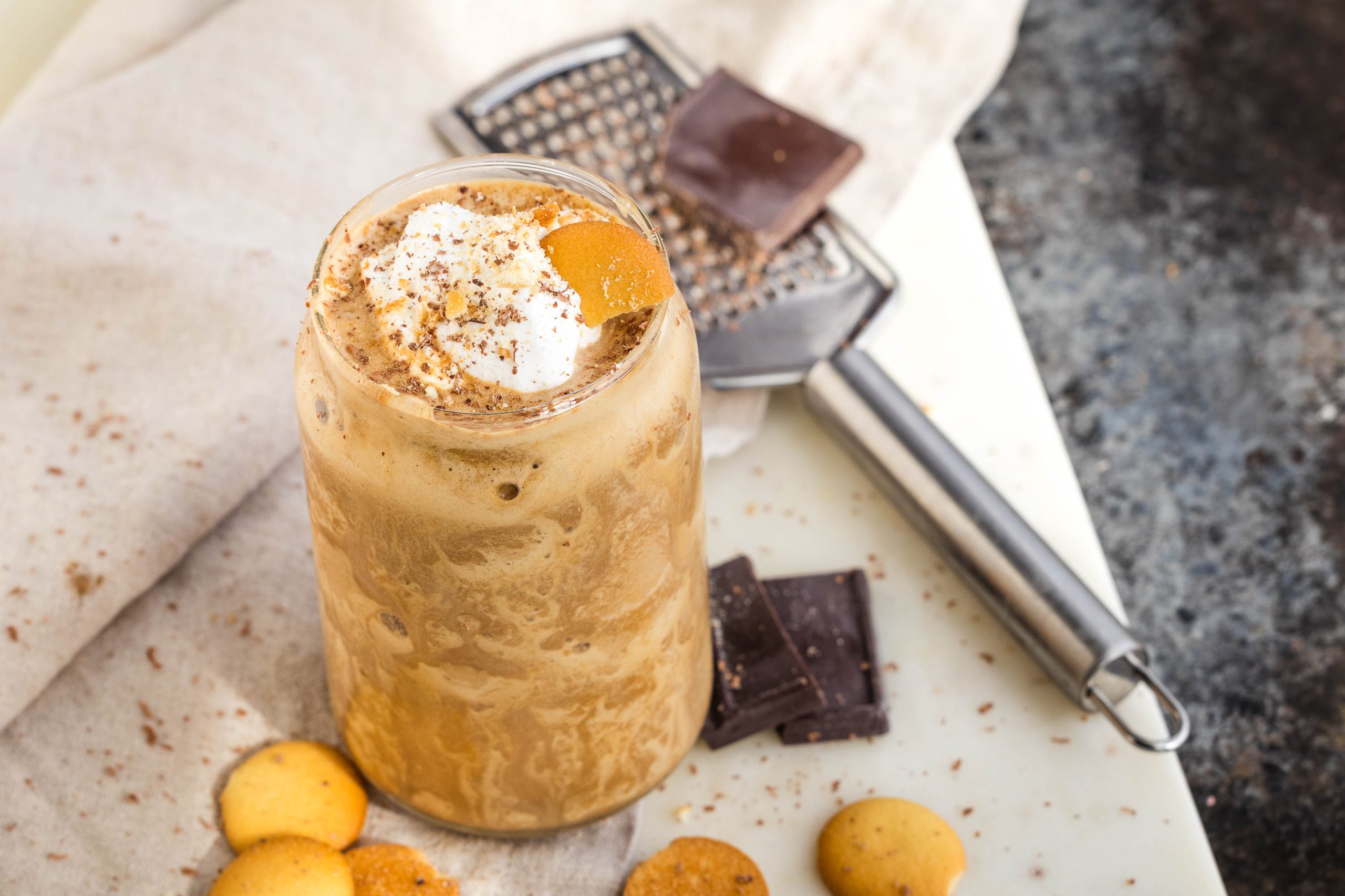 A tall glass of frothy Protein Tiramisu Smoothiewith whipped cream, chocolate shavings, and an orange slice garnish, surrounded by chocolate pieces, a grater, and small cookies on a cloth.