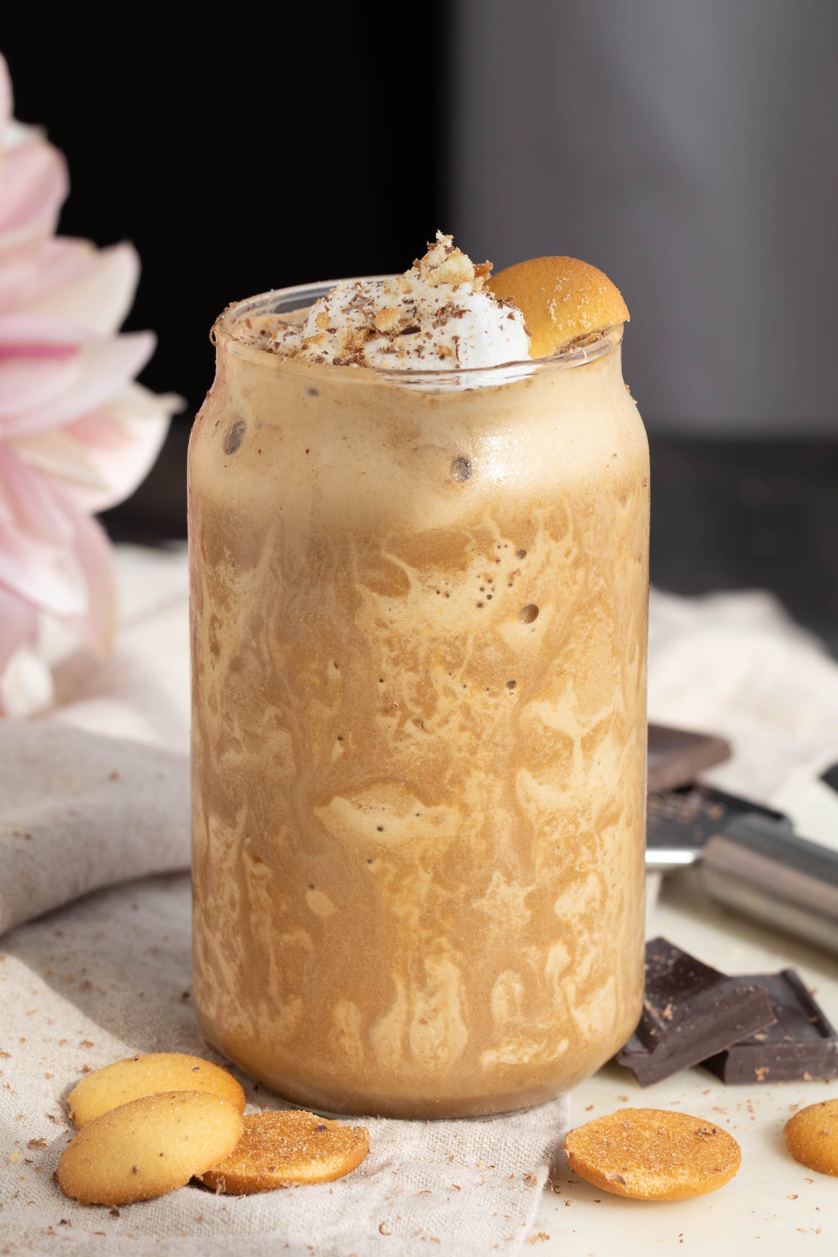 A frothy Tiramisu smoothie in a glass jar topped with whipped cream, cocoa powder, and a cookie. Cookies are placed around the jar on a cloth, with chocolate pieces nearby. A pink flower peeks in from the side.