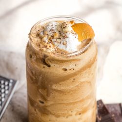 A creamy, frothy coffee drink in a glass, topped with whipped cream, a peach slice, and a sprinkle of spice. Surrounded by chocolate pieces and small round cookies.