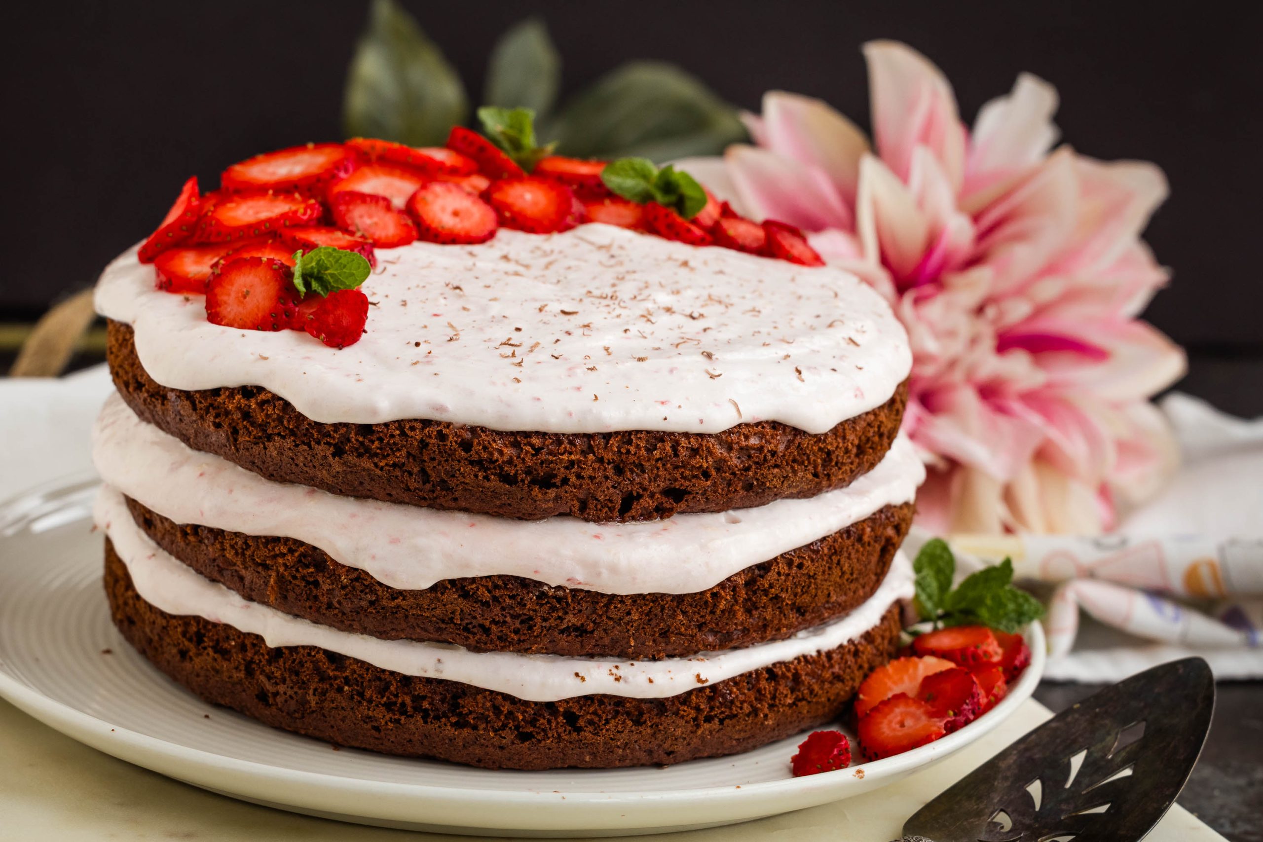 A three-layer chocolate cake with white frosting and sliced strawberries on top, garnished with mint leaves, on a white plate. A large pink flower is in the background.