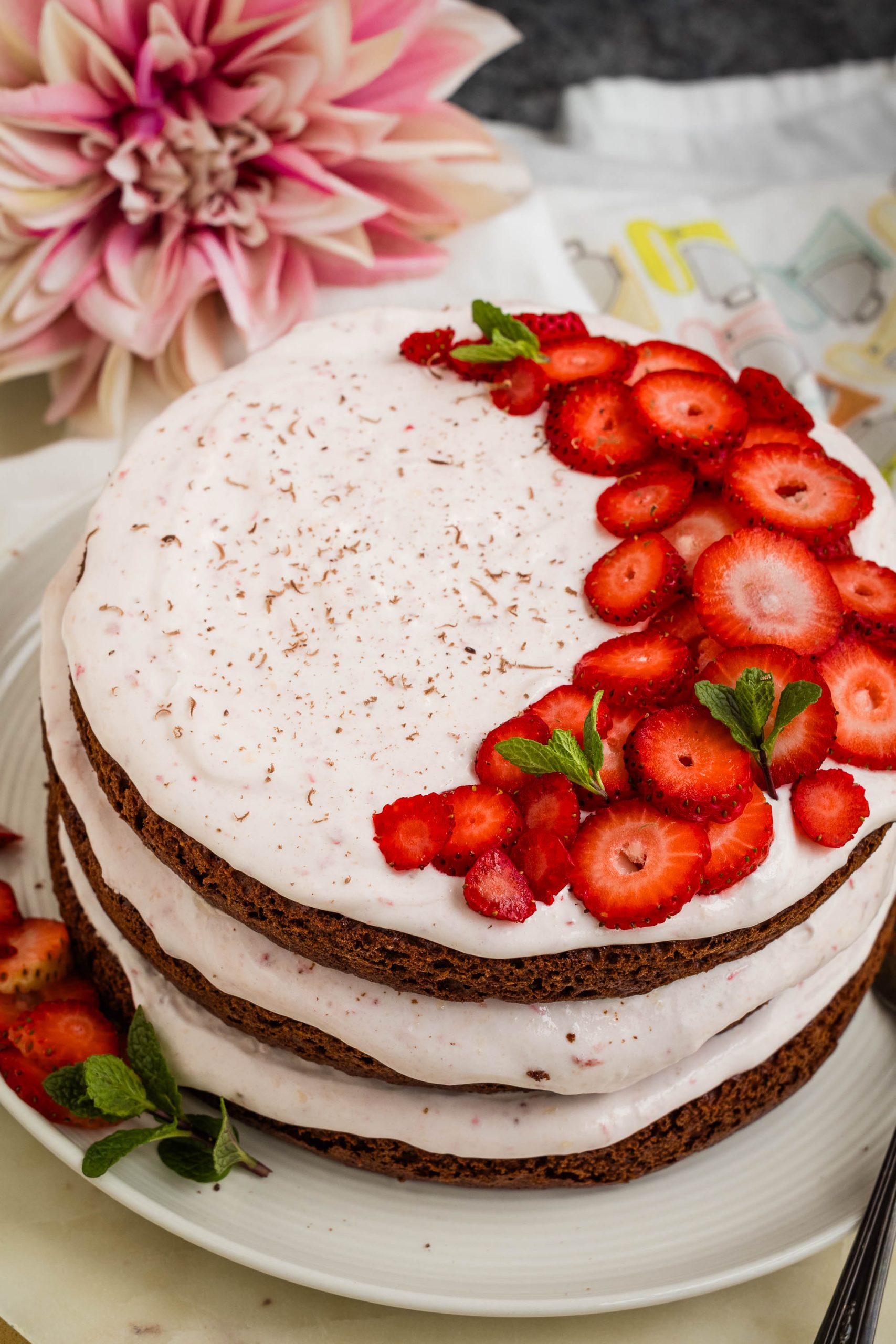 A layered chocolate cake with white frosting, garnished with sliced strawberries and mint leaves on top and sides, next to a large pink and white flower.