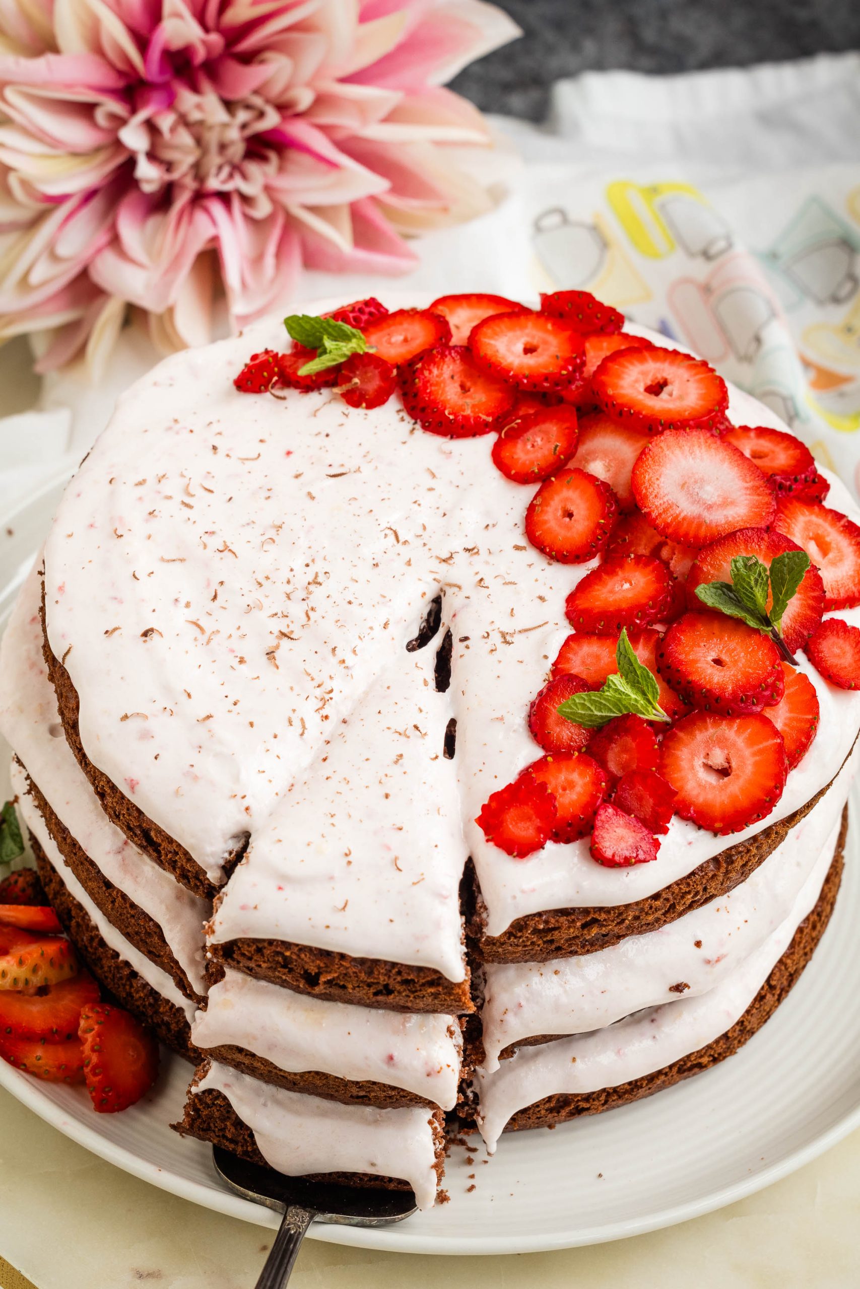 A layered strawberry chocolate cake with light frosting, topped with sliced strawberries and mint leaves. One slice is cut out and placed on the side. A large flower is in the background.