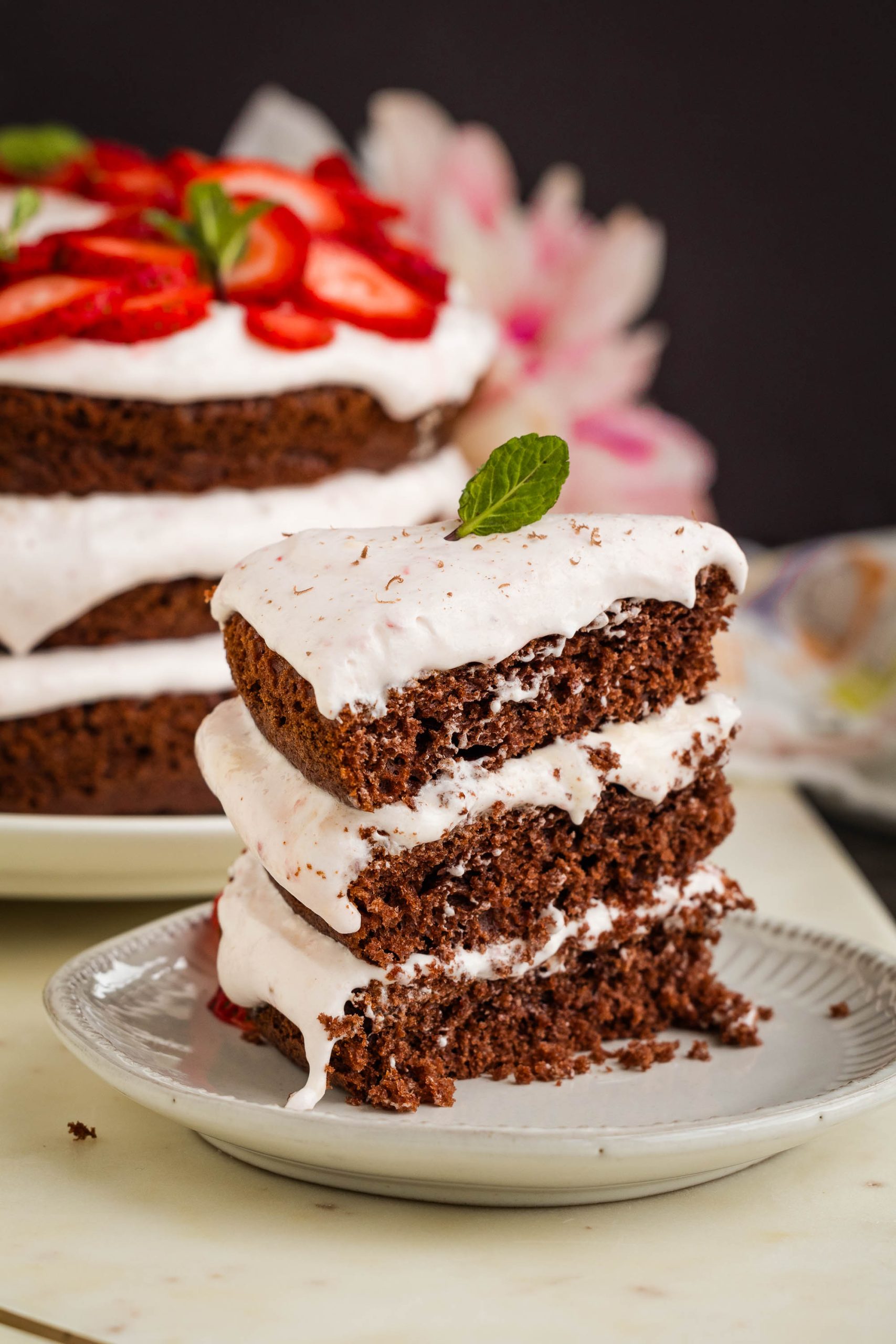 A slice of dessert with layers of white frosting, topped with a mint leaf, sits on a plate. Whole cake in background with sliced strawberries and mint garnish.