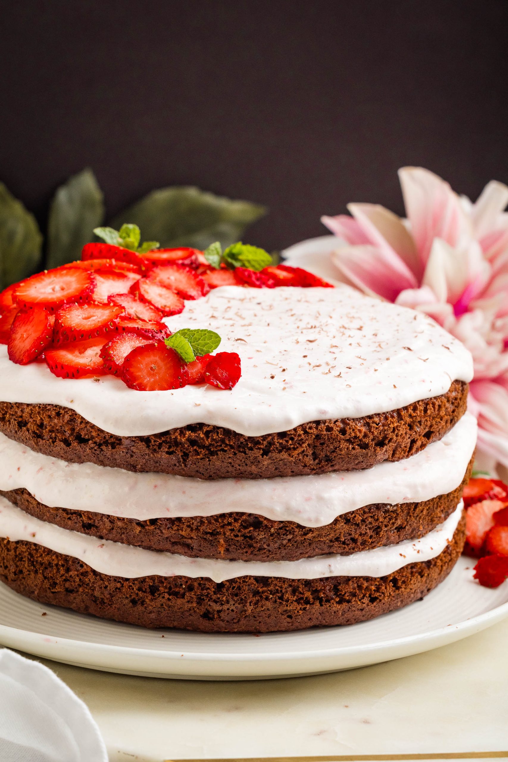 Three-layer strawberry chocolate cake with white frosting between layers, topped with sliced strawberries and mint leaves on a white plate.