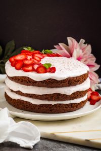 Three-layer chocolate cake with white frosting, topped with sliced strawberries and mint leaves. Placed on a white plate, a pink flower is visible in the background.