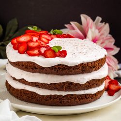 Three-layer chocolate cake with white frosting, topped with sliced strawberries and mint leaves. Placed on a white plate, a pink flower is visible in the background.