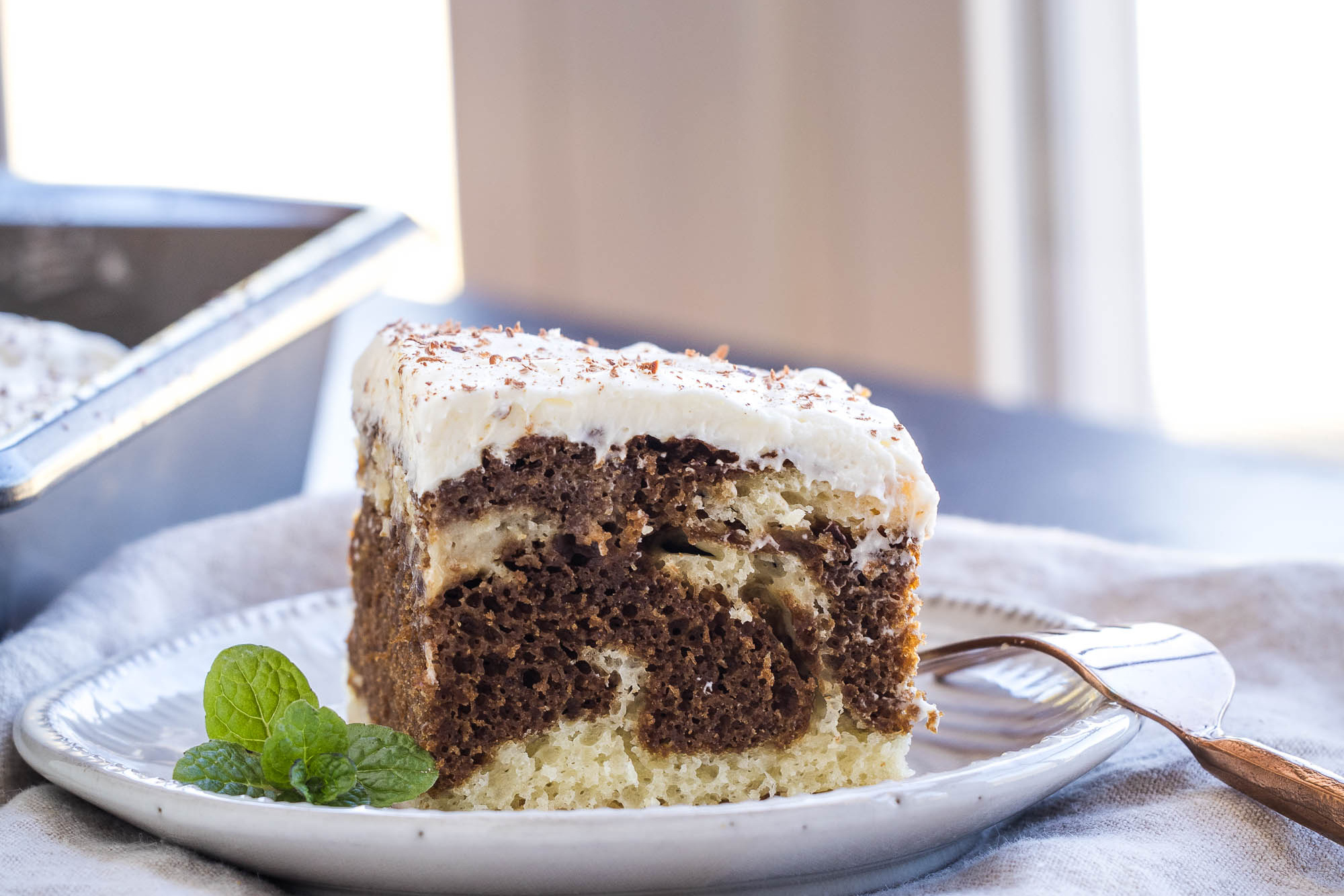 A slice of marble cake with white frosting sits elegantly on a plate, garnished with mint leaves. Beside it, a fork invites you to indulge, reminiscent of a classic tiramisu.