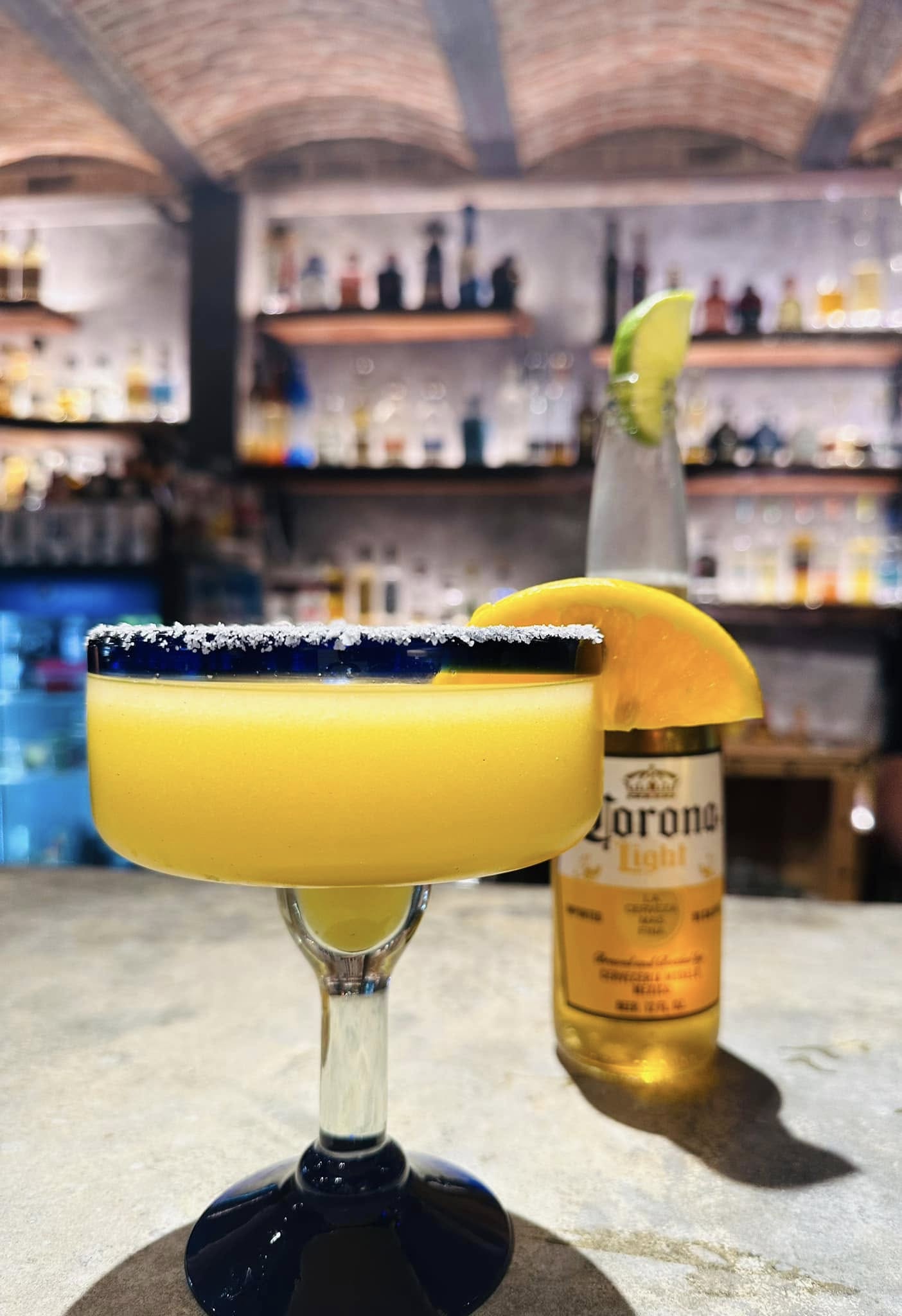 A margarita with a salted rim is in the foreground, and a Corona Light beer with a lime slice is in the background, set on a bar counter.