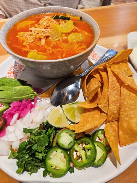 Cantina Del Sol. A bowl of chicken soup with yellow squash and a plate of garnishes including avocado, lime, cilantro, onions, sliced jalapeños, pink pickled onions, and tortilla chips.