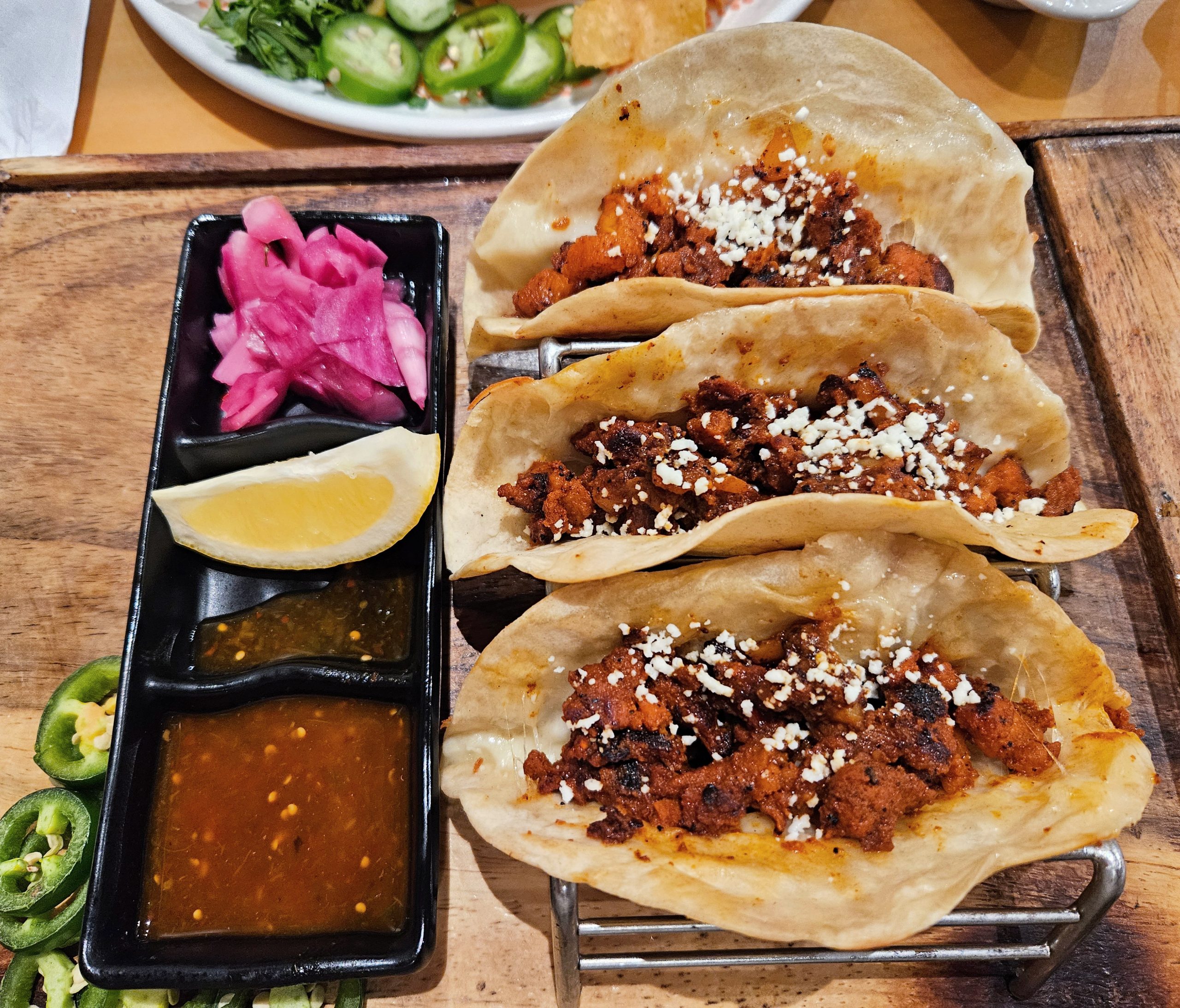 Cantina Del Sol. Three tacos filled with seasoned meat topped with crumbled cheese on a wooden platter. Pickled onions, lemon wedge, and two types of sauce are in a tray alongside. Sliced jalapeños visible.