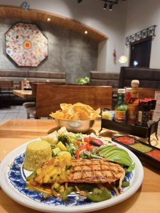 A plate of grilled chicken with mango salsa, rice, avocado, and vegetables is on a table. A basket of tortilla chips and a condiment caddy are in the background within a restaurant setting.