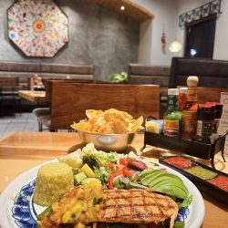 A plate of grilled chicken with mango salsa, rice, avocado, and vegetables is on a table. A basket of tortilla chips and a condiment caddy are in the background within a restaurant setting.