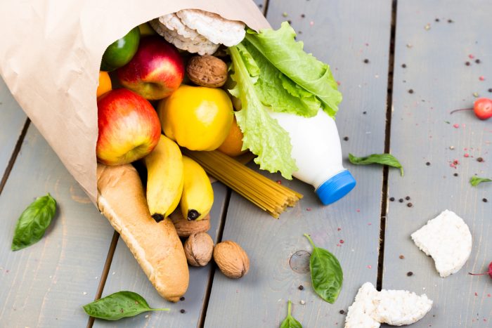 A paper bag overflows with groceries, including apples, bananas, a baguette, spaghetti, lettuce, walnuts, rice cakes, a milk bottle, and various leaves, scattered on a wooden surface.