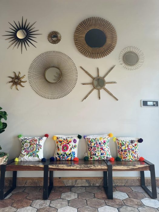 Cantina Del SolWall with decorative sunburst mirrors above a bench featuring four colorful pillows with floral designs and tassels. Hexagonal floor tiles and a small plant on the left.