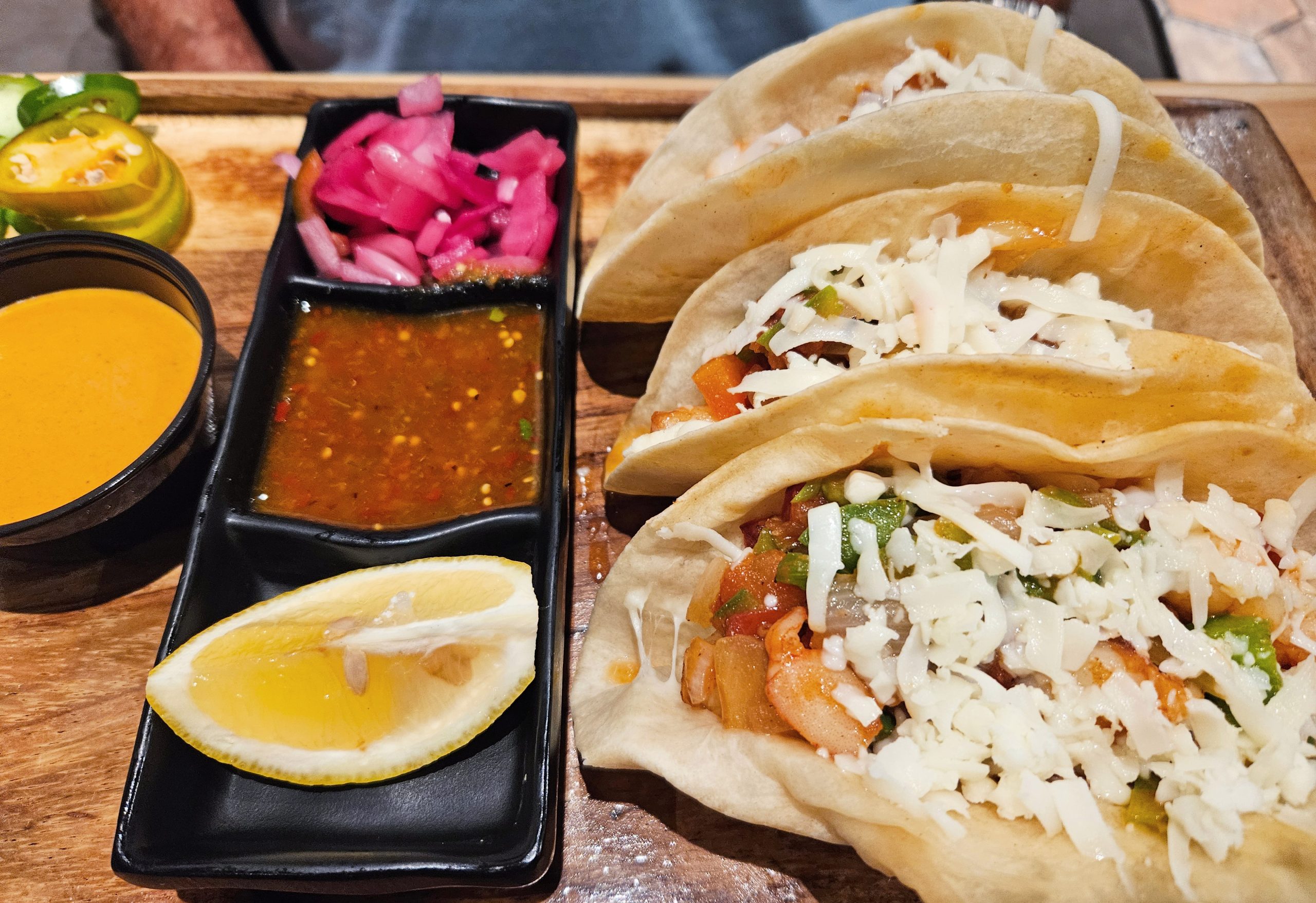 Three tacos with cheese, served with red and yellow sauces, pickled onions, sliced jalapeños, and a lemon wedge on a wooden tray.